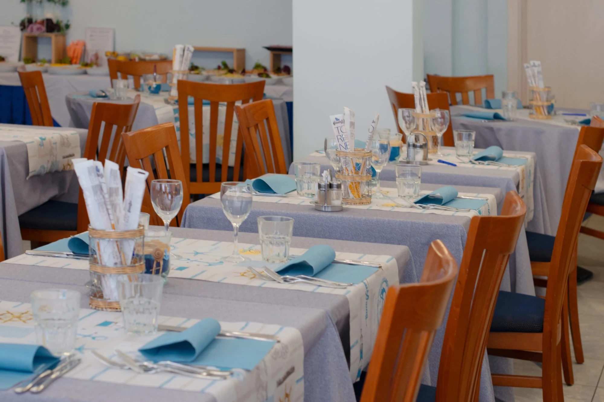 Sala ristorante con tavoli apparecchiati, tovaglie azzurre e bianche, bicchieri e tovaglioli blu