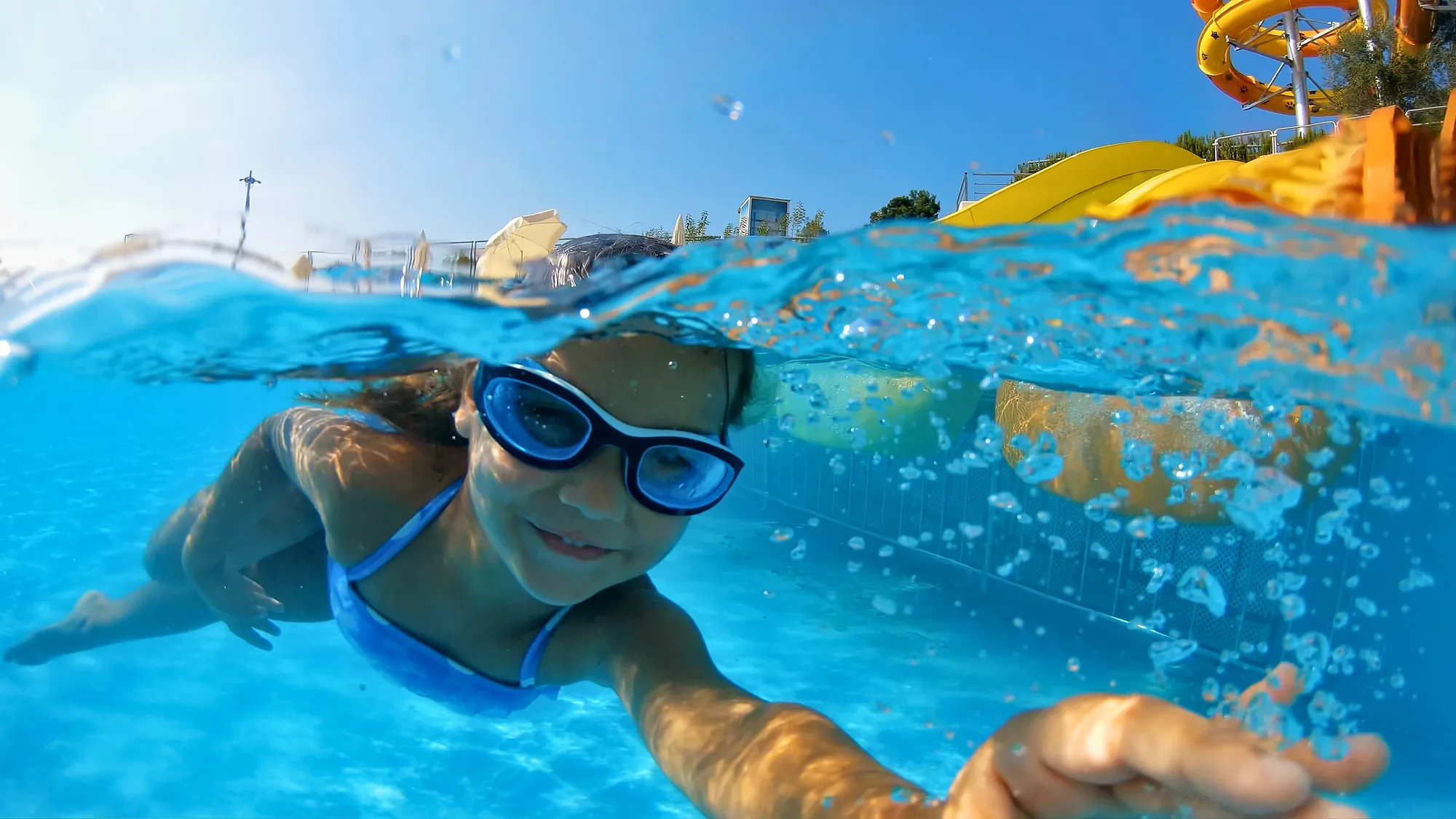 Una bambina con gli occhialini nuota a pelo d’acqua in un parco acquatico, con gli scivoli alle spalle