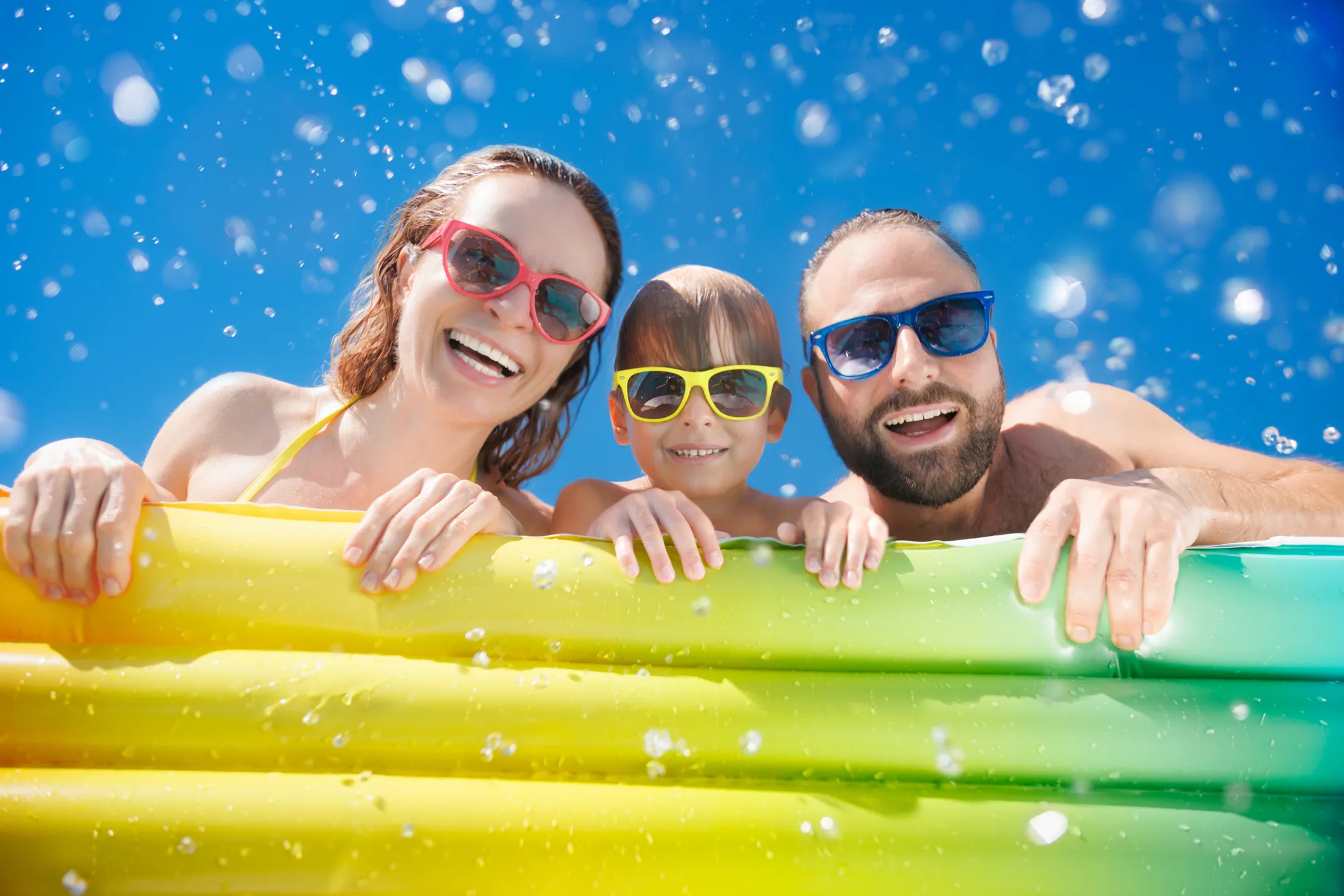 Una famiglia con gli occhiali da sole sorride appoggiata a un materassino giallo in piscina
