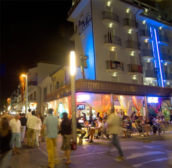 Hotel illuminato di notte con persone a passeggio e clienti seduti al bar all'aperto