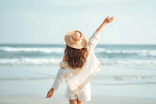 Donna di spalle sulla spiaggia che osserva il mare, con un cappello di paglia e un abito leggero bianco, mentre alza un braccio verso il cielo in una giornata luminosa e rilassante.