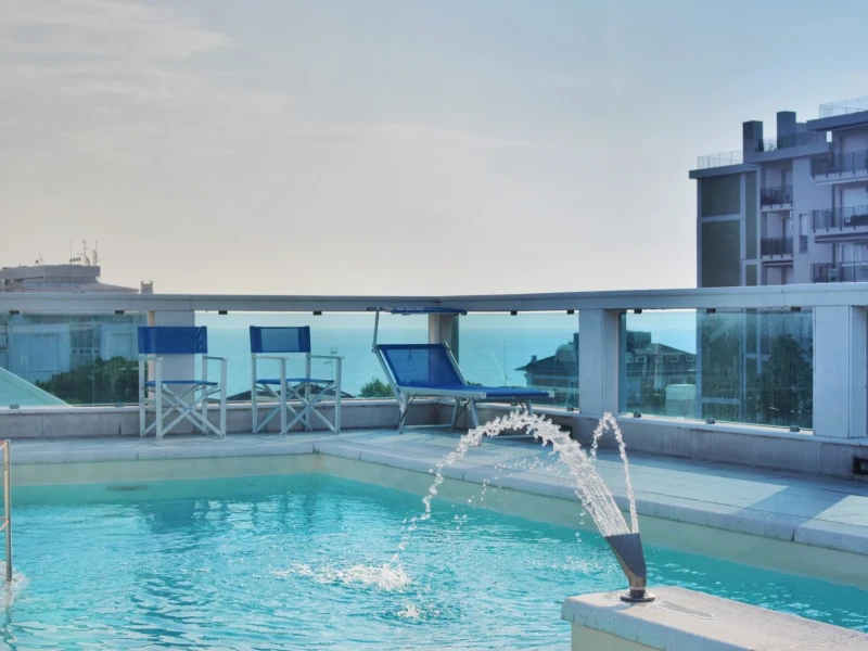 A rooftop pool with a water fountain and blue lounge chairs overlooking the city and sea.