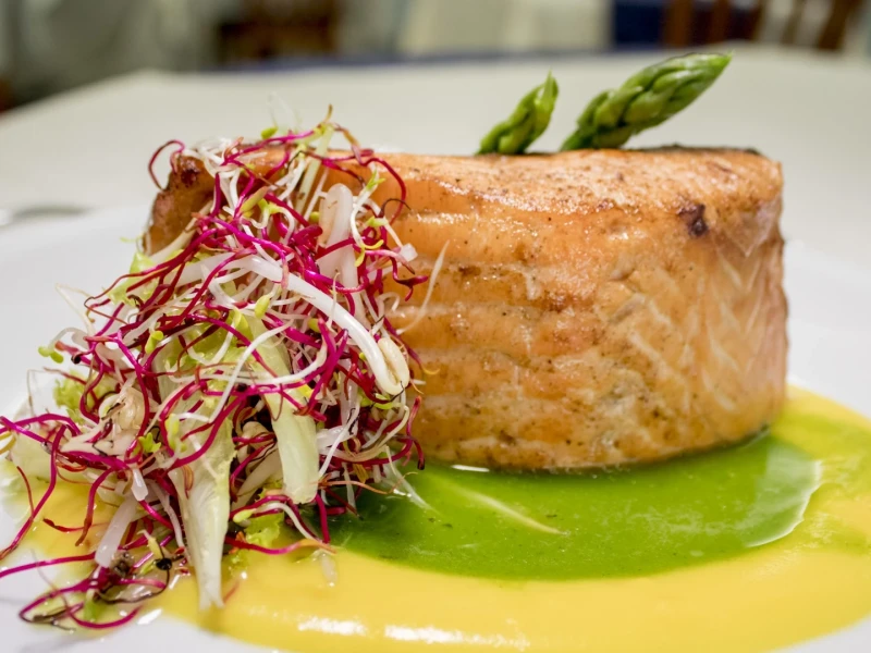 A dish featuring grilled salmon with colorful sprouts and asparagus on a bed of yellow and green sauces.