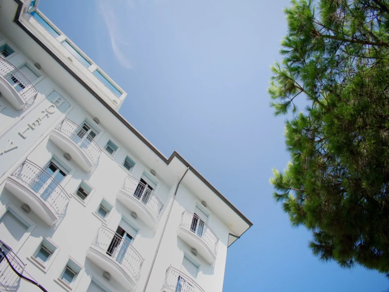 A white building with balconies next to a green tree against a clear blue sky.
